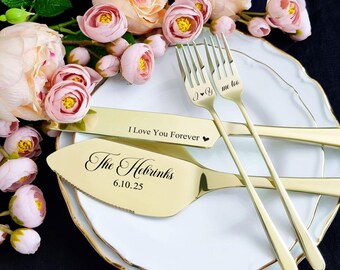 Könnte beinhalten: Goldfarbenes Hochzeits-Kuchen-Servierset mit den Gravuren "I Love You Forever" und "The Hebrinks 6.10.25". Das Set beinhaltet einen Tortenheber, ein Messer und zwei Gabeln. Das Set liegt auf einem weißen Teller mit Goldrand, umgeben von rosa Blumen.