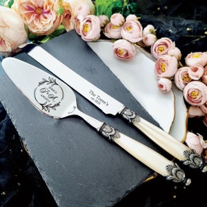 May include: A silver cake knife and server set with white pearl handles and ornate silver accents. The knife is engraved with "The Torre's est. 2023" and the server is engraved with "Jessica & Angelo" in a floral wreath.