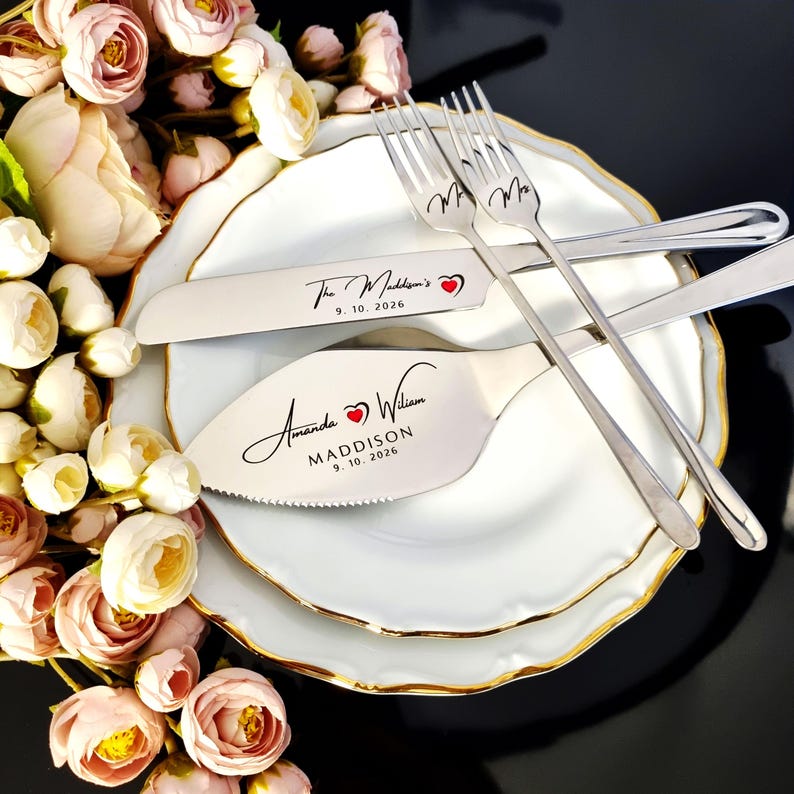 Personalized 4-piece silver wedding cake cutting set with a red heart engraved detail. Mirror-polished stainless steel knife, server, and Mr. and Mrs. forks for the Maddison wedding, 9.10.2026. Styled on gold-rimmed plates with pink roses.