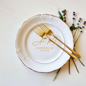 May include: A white ceramic plate with a gold rim and embossed edge, featuring the gold text GONZALEZ 07.03.26. Two gold forks are crossed on the plate, each engraved with the date 07.03.26. A sprig of purple lavender rests beside the plate.