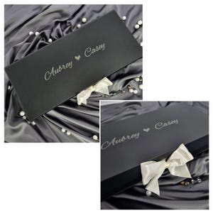 Personalized premium black magnetic wedding gift box with a white satin ribbon bow. Custom engraved with Aubrey and Casey in elegant script. Staged on luxury charcoal grey silk fabric with scattered decorative pearls for a gift-ready look.