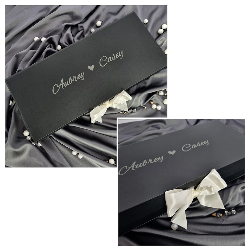 Personalized premium black magnetic wedding gift box with a white satin ribbon bow. Custom engraved with Aubrey and Casey in elegant script. Staged on luxury charcoal grey silk fabric with scattered decorative pearls for a gift-ready look.