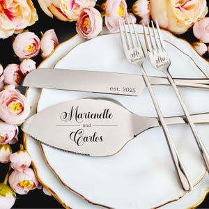 Personalized 4-piece silver wedding cake cutting set for the Rowald wedding. Server engraved with names Marienelle and Carlos; knife features est. 2025. Set includes Mr. Rowald and Mrs. Rowald forks, styled on a gold-rimmed plate with pink roses.