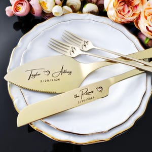 Personalized 4-piece champagne gold wedding cake cutting set with Swarovski heart crystal inlays. Engraved with names and date in modern script. Set includes knife, server, and two dessert forks, shown on a white plate with romantic roses.