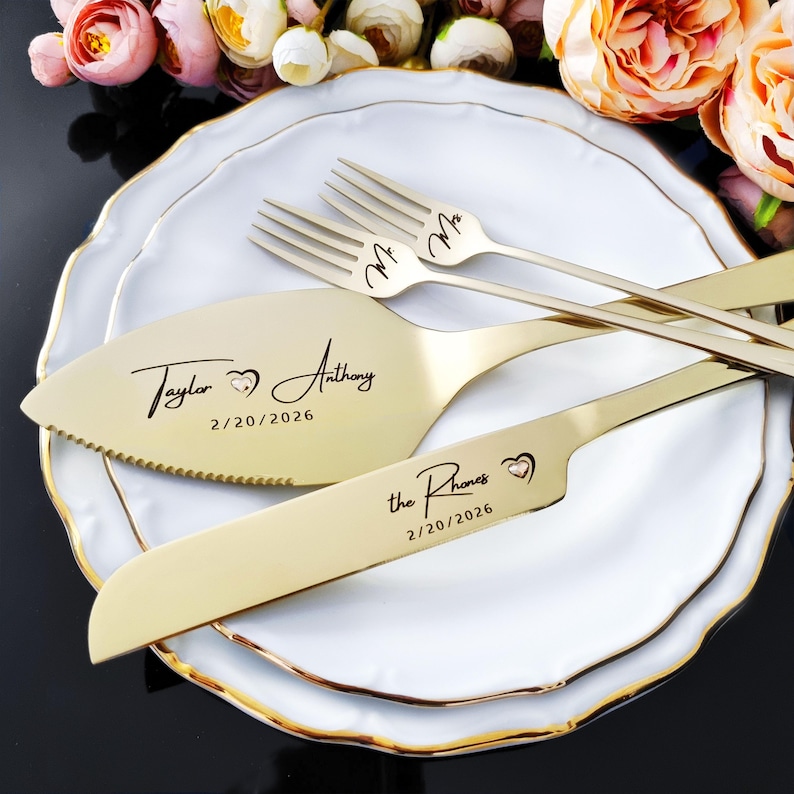 Personalized 4-piece champagne gold wedding cake cutting set with Swarovski heart crystal inlays. Engraved with names and date in modern script. Set includes knife, server, and two dessert forks, shown on a white plate with romantic roses.