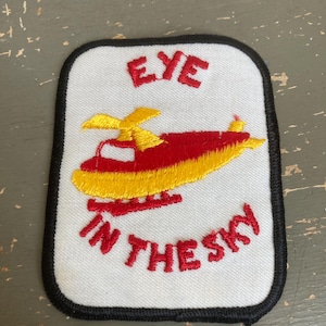 May include: Embroidered patch with a white background, black border, and a red and yellow helicopter design. The text "EYE" is above the helicopter, and "IN THE SKY" is below.