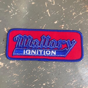 May include: A rectangular patch with a red background and a blue border. The patch features the word "Mallory" in a stylized blue font above the word "Ignition" in white, also in a blue border.