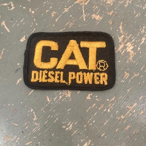 May include: Black rectangular patch with gold embroidered text reading "CAT DIESEL POWER". The patch features the Caterpillar logo. The patch is likely for a jacket or bag.