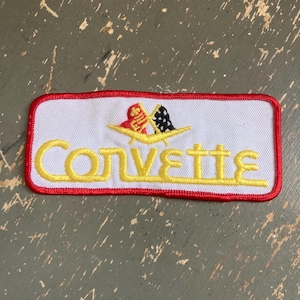 May include: Embroidered patch with the word "Corvette" in yellow letters on a white background. The patch has a red border and features a logo with two flags. The patch is rectangular in shape.