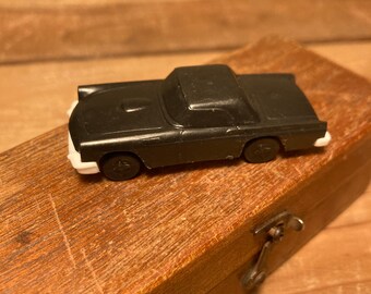May include: A black toy car in a wooden box. The car has white trim around the wheels and a black body.
