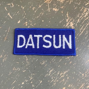 May include: A rectangular embroidered patch featuring a deep blue background with the word DATSUN stitched in bold white capital letters. The patch has a finished blue border and a flat, textured surface consistent with standard fabric embroidery.
