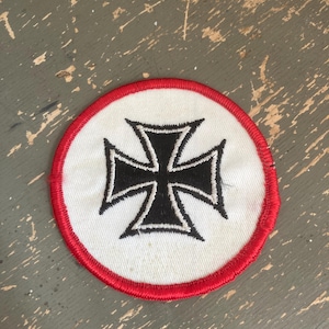 May include: Round patch featuring a black cross design on a white background, encircled by a red border. The patch is likely made of fabric and is suitable for sewing or ironing onto clothing or accessories.