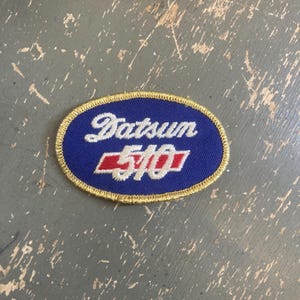 May include: An oval-shaped embroidered patch featuring a blue background with a gold-colored stitched border. The text Datsun is embroidered in white script above the number 510, which is displayed in white font over a horizontal red rectangular bar.