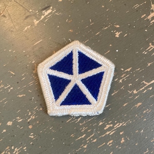 May include: A blue and white embroidered patch in a pentagon shape. The patch features a blue star design on a white background, with a white border. The patch is likely a military or organizational emblem.