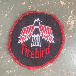 May include: Round black patch with a red stitched border. The patch features a white and red bird design with the word "Firebird" embroidered in red. The patch is likely for clothing or accessories.