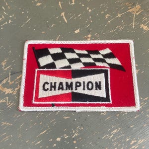 May include: A rectangular patch with a red background, white border, and the word "CHAMPION" in a white and black design. A black and white checkered flag is above the word. The patch is likely for a jacket or other apparel.