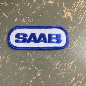 May include: A rectangular patch with the word "SAAB" embroidered in blue on a white background. The patch has a blue border and is likely an emblem or logo.