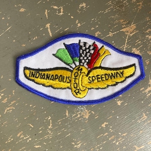 May include: Embroidered patch with a white background, blue border, and gold wings. The patch reads "Indianapolis Speedway" in gold lettering. Above the wings are green, blue, and red flags, and a checkered flag.