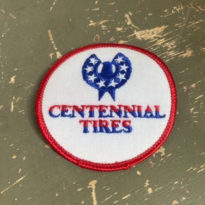 May include: A round patch with a white background and a red border. The patch features a blue design with stars and the words "CENTENNIAL TIRES" in blue.