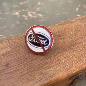 May include: A round pin with a white border, red outline, and a blue and white Ford logo with a red slash through it. The pin is on a wooden surface.