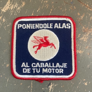 May include: A square patch with a red border, navy blue center, and white text. The text reads "PONIENDOLE ALAS" and "AL CABALLAJE DE TU MOTOR". A red winged horse is in the center.