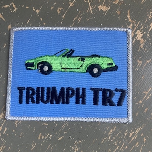 May include: A rectangular patch with a light blue background and a green convertible car design. The text "TRIUMPH TR7" is embroidered in black. The patch has a silver border.