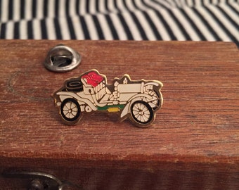May include: A gold-colored pin of a white vintage car with red seats and black tires. The car is on a wooden surface.