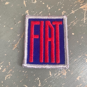 May include: A square patch featuring the word "FIAT" in bold red letters on a blue background. The patch has a silver border and is likely an emblem or logo.