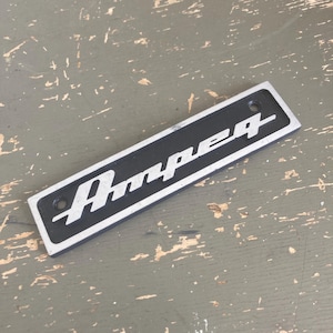 May include: A rectangular metal emblem featuring the word Ampeg in a stylized silver script font set against a black background. The emblem has a silver border and two small circular mounting holes, one on each end of the rectangular plate.