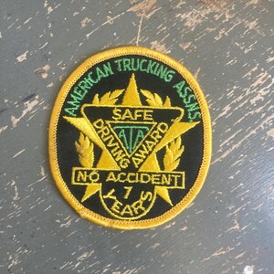 May include: A circular embroidered patch with a black background and yellow border. It features a yellow star, green text reading AMERICAN TRUCKING ASSNS, and a central triangle with the text SAFE DRIVING AWARD, NO ACCIDENT, and 7 YEARS.
