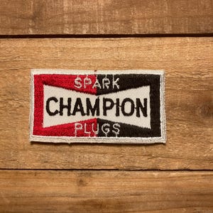 May include: A red, white, and black embroidered patch with the text "Spark Plug Champion" in white lettering.
