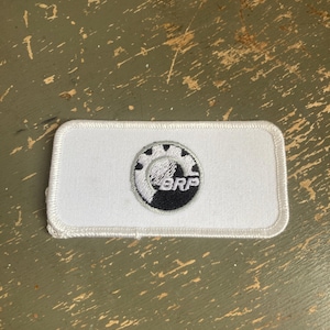 May include: White rectangular patch with a black and white circular logo featuring the letters "BRP". The patch has a white border and is likely an emblem or insignia.