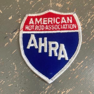 May include: A shield-shaped patch with a red top section and a blue bottom section. The text "AMERICAN HOT ROD ASSOCIATION" is in white on the red section. The letters "AHRA" are in white on the blue section. The patch has a white stitched border.