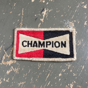 May include: Rectangular Champion patch with a red and black design. The patch features the word "CHAMPION" in bold, black letters against a cream-colored background. The patch is bordered with a cream-colored thread.