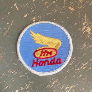 May include: A round patch featuring the Honda logo. The patch has a light blue background with a gold wing design and a red oval with the letters "HM" inside. The word "Honda" is embroidered in red.