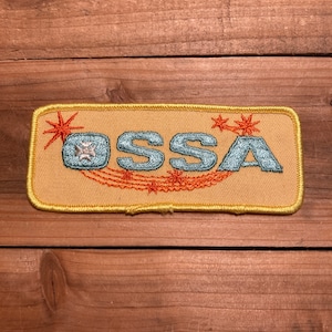 May include: A yellow embroidered patch with the word "OSSA" in blue and orange lettering. The patch has a yellow border and features a starburst design with orange and white stars.