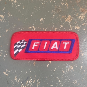 May include: A rectangular red patch featuring the word "FIAT" in white letters, outlined in blue. A checkered flag is on the left side. The patch is likely an emblem or logo.