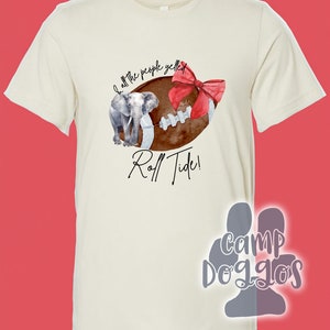May include: Cream-colored t-shirt featuring a watercolor design of a football with a red bow, an elephant, and the text "Roll Tide!". The shirt has a classic crew neck and short sleeves.