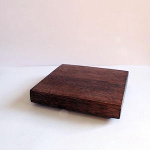 May include: A rectangular, dark brown wooden box with a flat top and a slightly raised base. The wood grain is visible, and the box appears to be made of a solid, sturdy material. The box is sitting on a white surface.