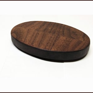May include: Oval-shaped wooden coaster with a dark brown, textured surface and a black edge. The coaster is made of wood and is designed to protect surfaces from drink condensation.