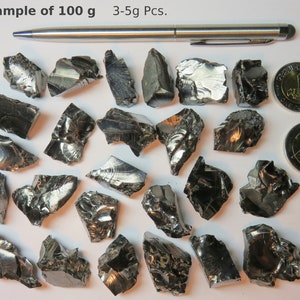 K&ouml;nnte beinhalten: Beispiel f&uuml;r 100 Gramm schwarzer, metallischer, unregelm&auml;&szlig;ig geformter St&uuml;cke. Die St&uuml;cke sind zwischen 3 und 5 Gramm schwer. Ein Stift und zwei Euro-M&uuml;nzen dienen als Gr&ouml;&szlig;envergleich.