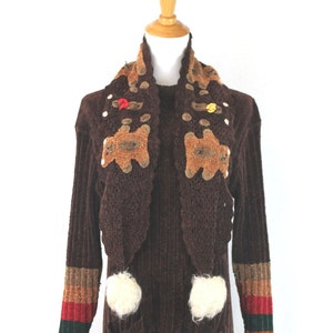 Puede incluir: Un suéter de punto marrón con una bufanda de ganchillo marrón con aplicaciones de ositos de peluche. El suéter tiene mangas largas con rayas de colores en los puños y pompones blancos en la parte delantera.