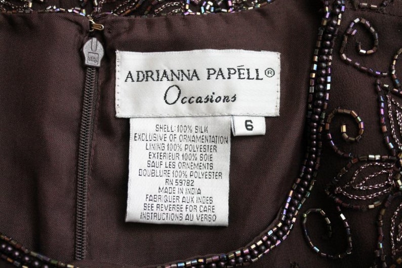 May include: A brown silk dress with beaded embellishments. The label reads "Adrianna Papell Occasions" and indicates the dress is size 6, made of 100% silk, and lined with 100% polyester.