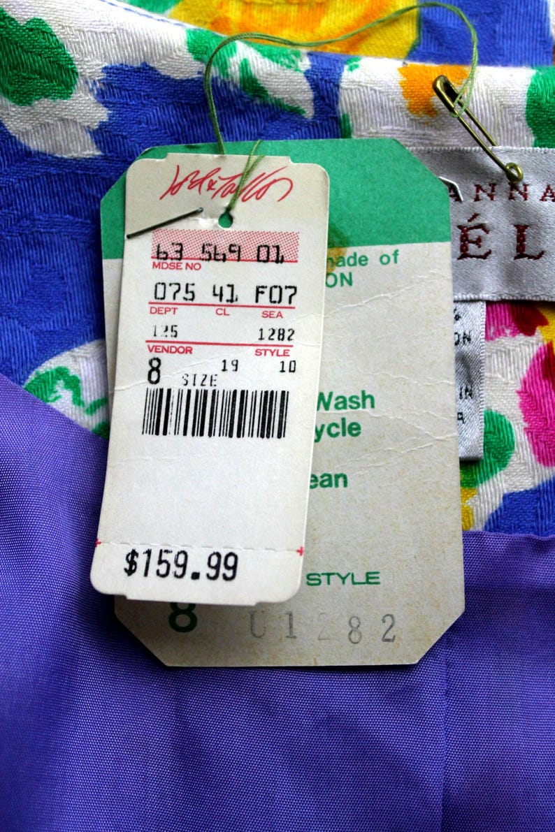 May include: A white, blue, green, and yellow floral fabric with a purple fabric underneath. A white tag with black text reads "63 569 01 MDSE NO 075 41 F07 DEPT CL SEA 125 1282 VENDOR STYLE 8 19 10 SIZE" and a barcode. The tag also has a price tag that reads "$159.99". A green tag with white text reads "made of ON Nash ycle ean STYLE 01282". A white tag with black text reads "ANNA &Eacute;L".