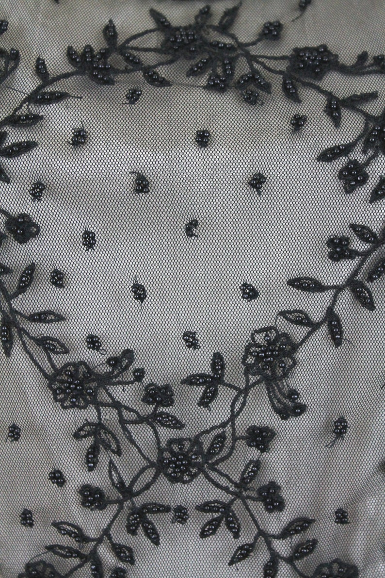 Pode incluir: Bordado floral preto com contas sobre um tecido preto transparente. O bordado apresenta delicados desenhos florais com pequenas contas pretas.