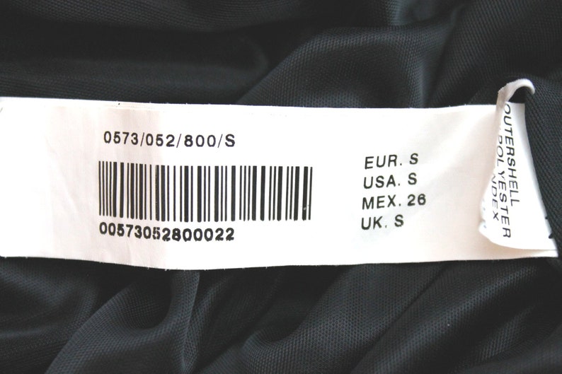 May include: A white tag with black text and a barcode. The tag shows the size of the garment in different sizing systems: EUR. S, USA. S, MEX. 26, UK. S. The tag also includes the product number 0573/052/800/S and the barcode number 00573052800022.