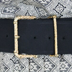 May include: A black fabric belt with a gold-toned, rope-like buckle. The belt is worn over a white and black patterned fabric.