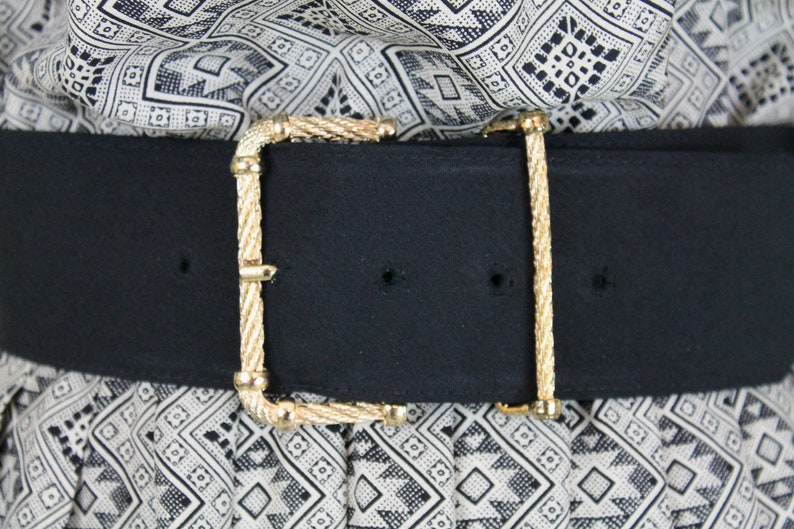 May include: A black fabric belt with a gold-toned, rope-like buckle. The belt is worn over a white and black patterned fabric.