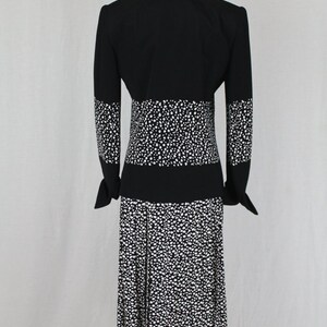 May include: A black and white polka dot dress with a black jacket. The dress has a fitted bodice and a skirt with pleats. The jacket has long sleeves and a collar.