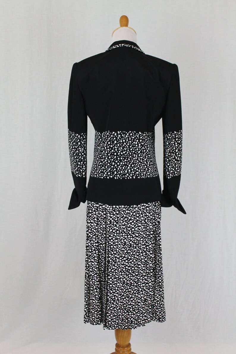 May include: A black and white polka dot dress with a black jacket. The dress has a fitted bodice and a skirt with pleats. The jacket has long sleeves and a collar.